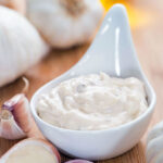 Serving of garlic aioli surrounded by garlic bulbs and cloves.
