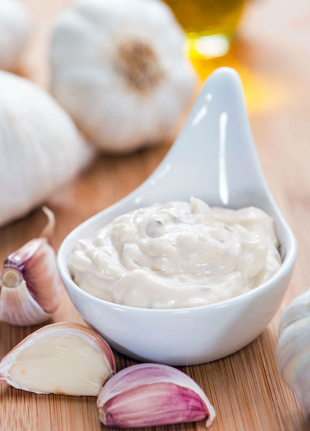 Serving of garlic aioli surrounded by garlic bulbs and cloves.