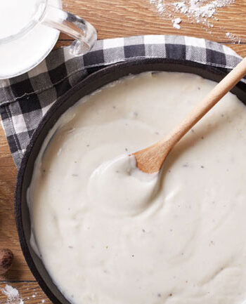 Pan of béchamel sauce and a wooden spoon with a checkered napkin and a pitcher of milk and flour on a cutting board.