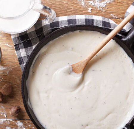Pan of béchamel sauce and a wooden spoon with a checkered napkin and a pitcher of milk and flour on a cutting board.