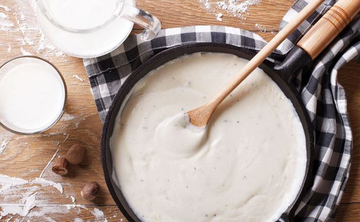 Pan of béchamel sauce and a wooden spoon with a checkered napkin and a pitcher of milk and flour on a cutting board.