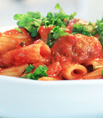 White bowl filled with rigatoni and meatballs and parsley garnish.