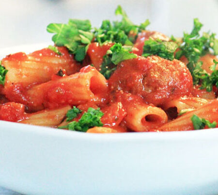 White bowl filled with rigatoni and meatballs and parsley garnish.