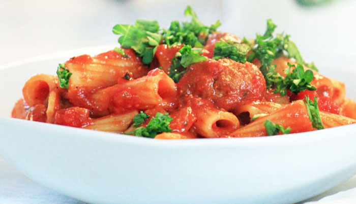 White bowl filled with rigatoni and meatballs and parsley garnish.