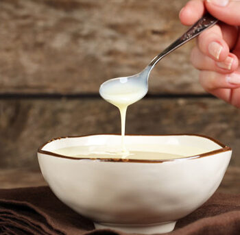 Bowl of sweetened condensed milk with a person holding a spoon and drizzling milk into the bowl.