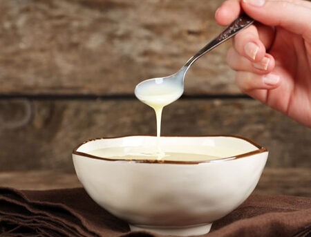 Bowl of sweetened condensed milk with a person holding a spoon and drizzling milk into the bowl.