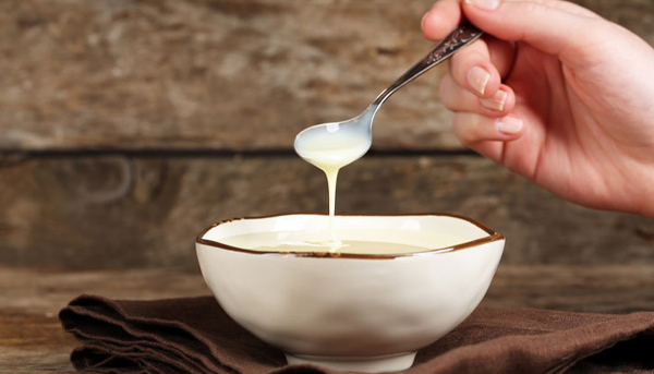 Bowl of sweetened condensed milk with a person holding a spoon and drizzling milk into the bowl.