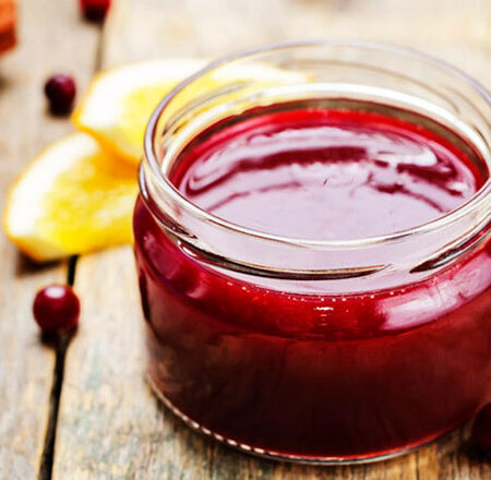 Open jar of Cranberry Strawberry Holiday Jam on a wood base with orange slices and cranberries.
