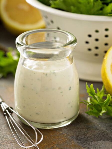 Vegan Old Bay Ranch Dressing in a glass bottle with a whisk, cut lemon, and a salad in a colander.