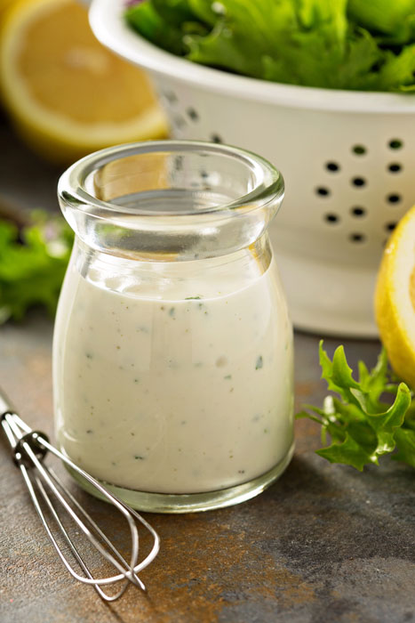 Vegan Old Bay Ranch Dressing in a glass bottle with a whisk, cut lemon, and a salad in a colander.