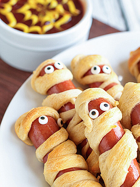 Vegan Halloween Mummy Dogs on a white plate with a ramekin filled with ketchup and mustard.