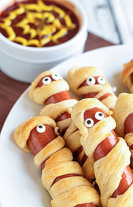 Vegan Halloween Mummy Dogs on a white plate with a ramekin filled with ketchup and mustard.